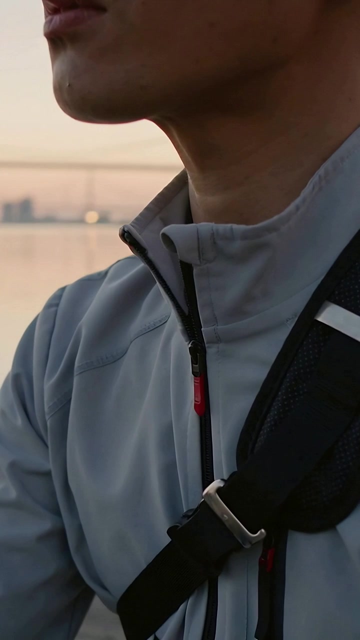 Vertical video: Adjusting jaw, man reacting to sunset while shifting jacket zip and strap at harbor