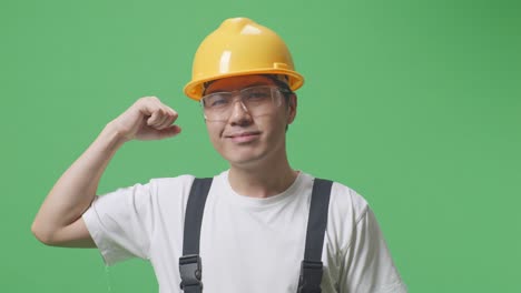 close up of asian man worker wearing goggles and safety helmet smiling and flexing his bicep while standing in the green screen background studio