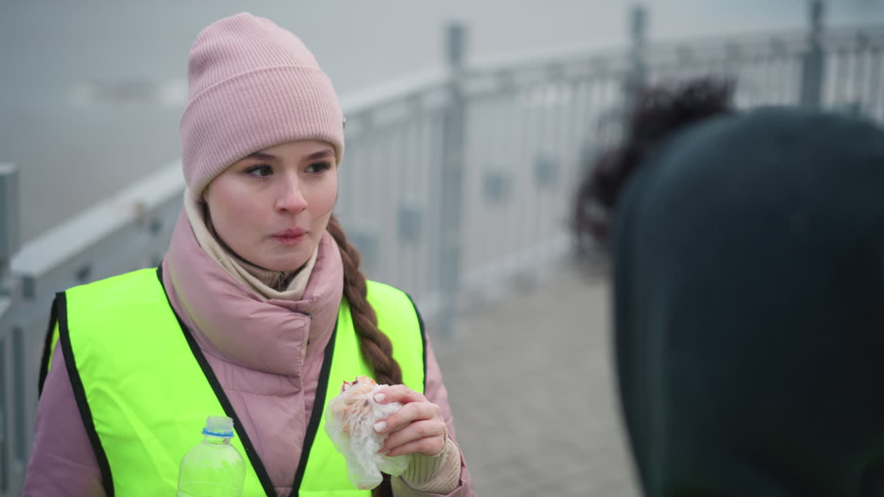 Smiling female worker wearing pink knit hat and reflective vest holding sandwich and bottle of water, standing outdoors near railing on cold day, looking down with shy happy expression during casual break