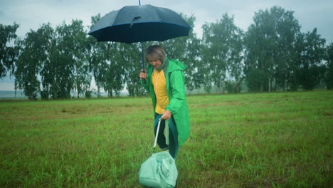 woman holds an open umbrella in one hand and bag in the other, walking in a grassy field, she drops the bag on the ground while, with a background of tall trees and a vast field