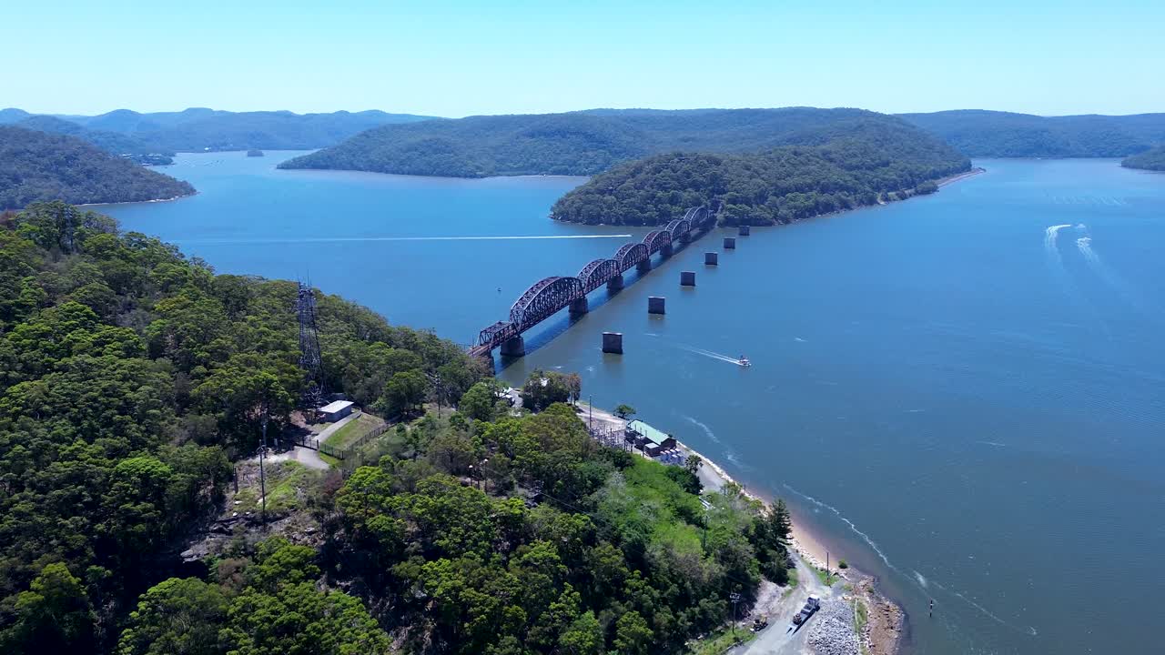 Paesaggio aereo con drone di foresta di boscaglia che copre un promontorio rurale con pescherecci che attraversano un canale dell'insenatura presso il ponte ferroviario sul fiume Hawkesbury a Brooklyn, Australia. Viaggi, turismo e natura.