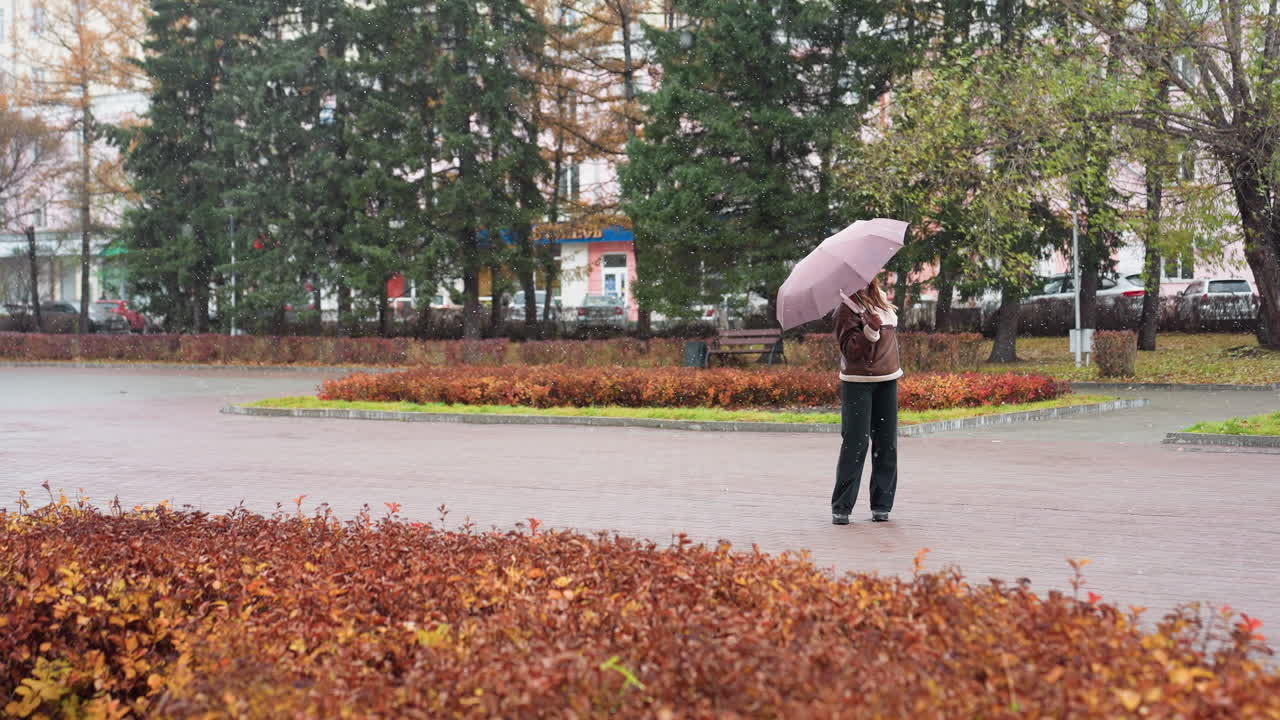 Cheerful girl holding umbrella, wearing knit cap, brown shearling jacket, black trousers, happily turning around in light snowfall, enjoying winter day, spreading positive energy in cold weather