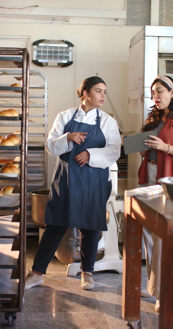 Chef della squadra femminile multietnica di una panetteria che indica uno scaffale mentre il manager tocca un tablet controllando il pane