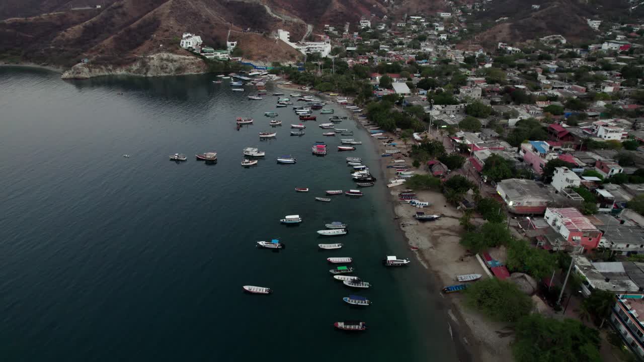 Cinematic boom shot lifting over the picturesque Taganga Bay, Colombia. Features a dense cluster of fishing boats and the coastal village nestled against the rugged Sierra Nevada mountains