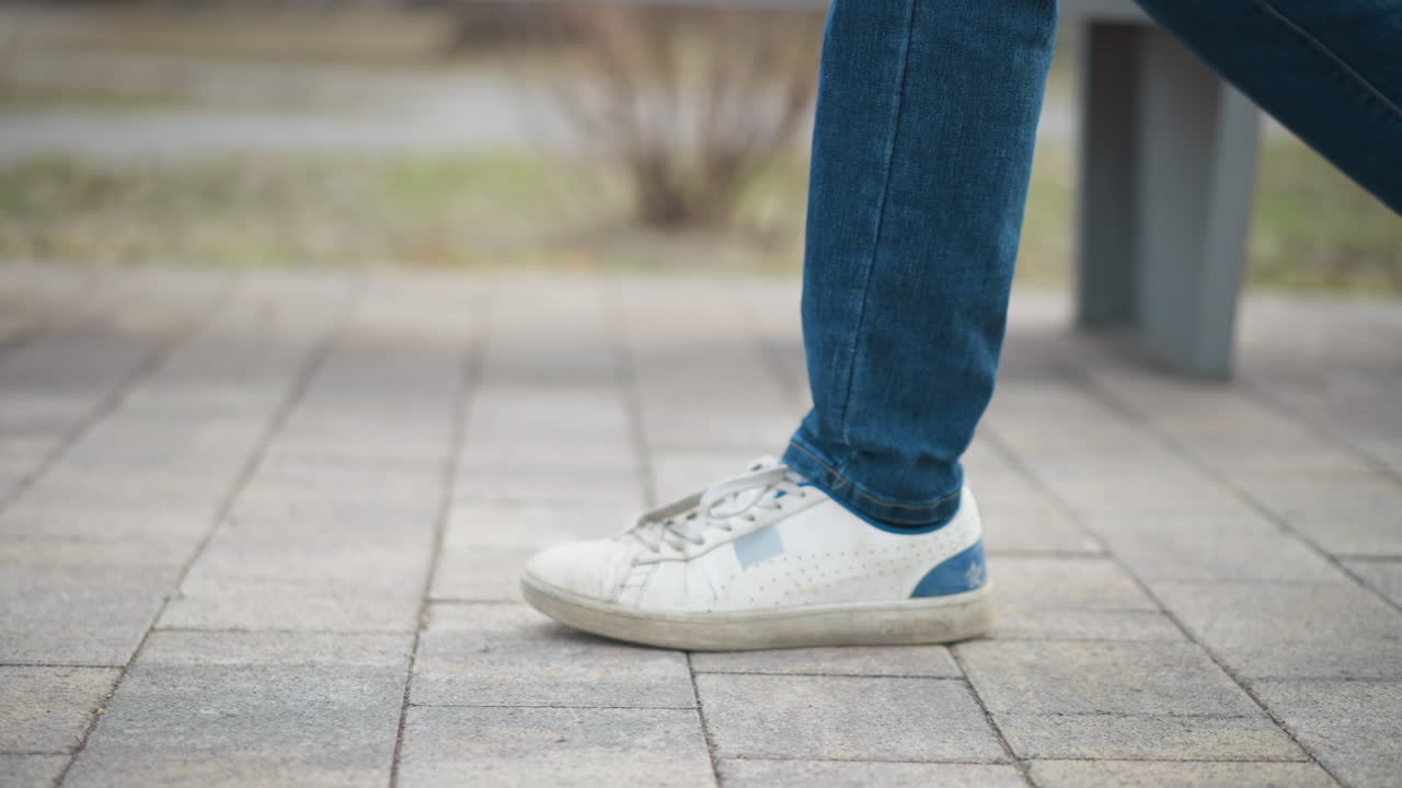 Black guy wearing jeans and white sneakers walking outdoors on paved path during early spring, close-up of both feet in motion, blurred grassy park background, casual urban style under soft cloudy daylight