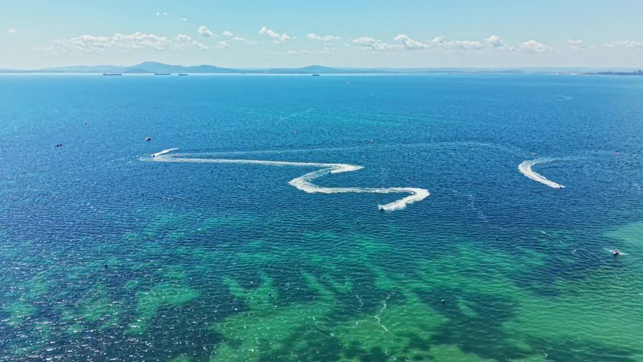 澄み切った空と遠くの丘が広がる晴れた日の沿岸地域で、広々とした水面を舞台にジェットスキーレースが行われる。