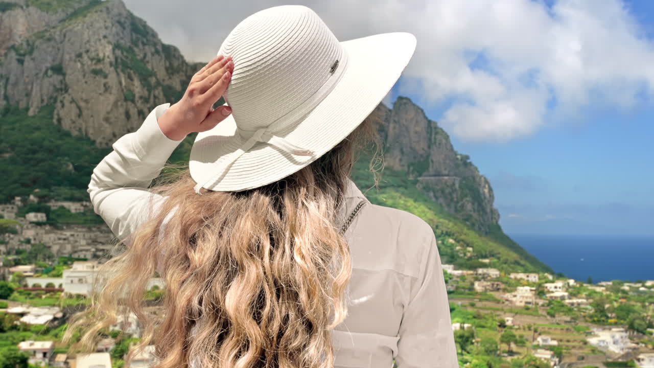 Woman in a white hat at a view point in Capri, Italy. View of the town, Tyrrhenian sea coast and greenery on the background