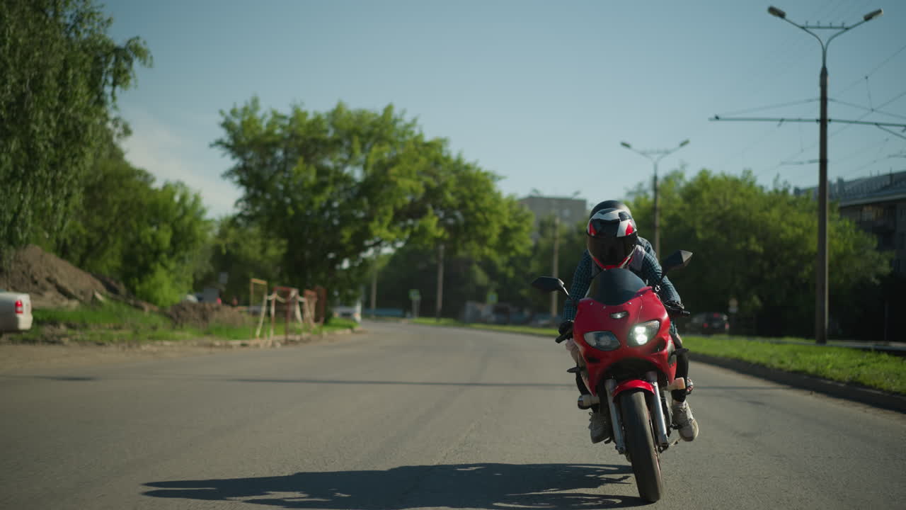 두 친구가 빨간색 전력 자전거를 타고 헬을 착용하고 도로 에 주차된 자동차와 함께 도로를 항해합니다. 전기 기둥과 울창한 나무가 배경에 보입니다.