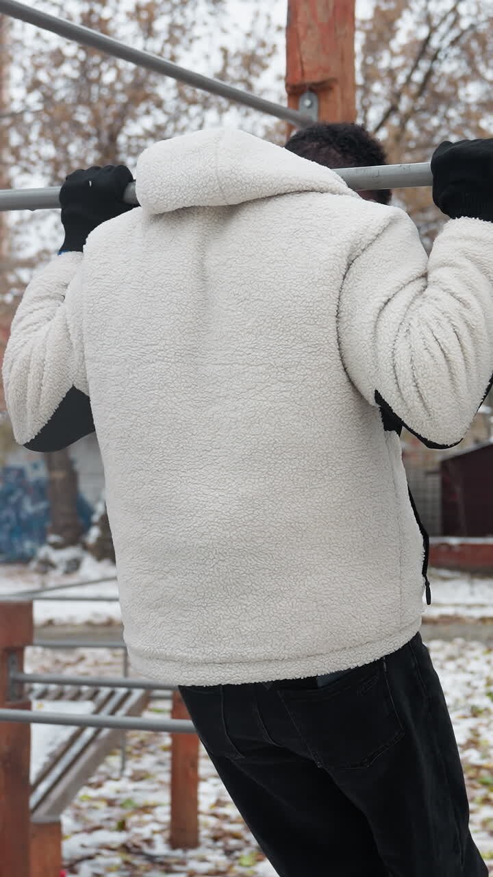 Student warms up for a workout, jumping to grab outdoor workout equipment, performing pull-ups and immediately dropping down in an urban setting with buildings, a car, and bare trees
