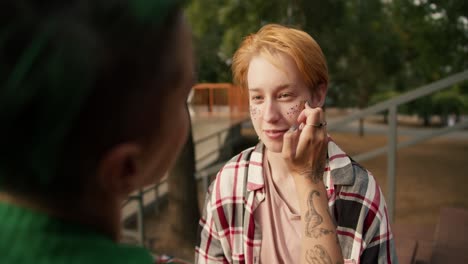 over the shoulder: a girl in a green shirt with short straight hair wears glitter yes, the cheeks of her girlfriend with short orange hair in a checkered pink shirt, lgbt couple in the park doing makeup