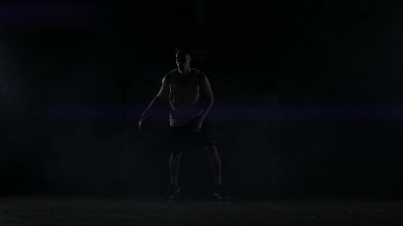 A basketball player dribbles down the court out of the shadows