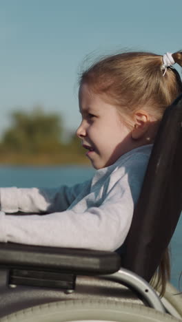 mother and daughter with paralysis walk near pond. blonde girl claps hands on armrests of wheelchair looking into distance on blurred background closeup