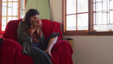 mujer de raza mixta relajada con auriculares sentada bebiendo té en la sala de estar de una cabaña soleada