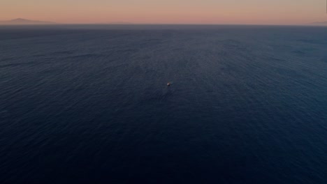 un solo barco flota solo en el medio de una gran extensión azul con un cielo de gradiente naranja rojo en la hora dorada