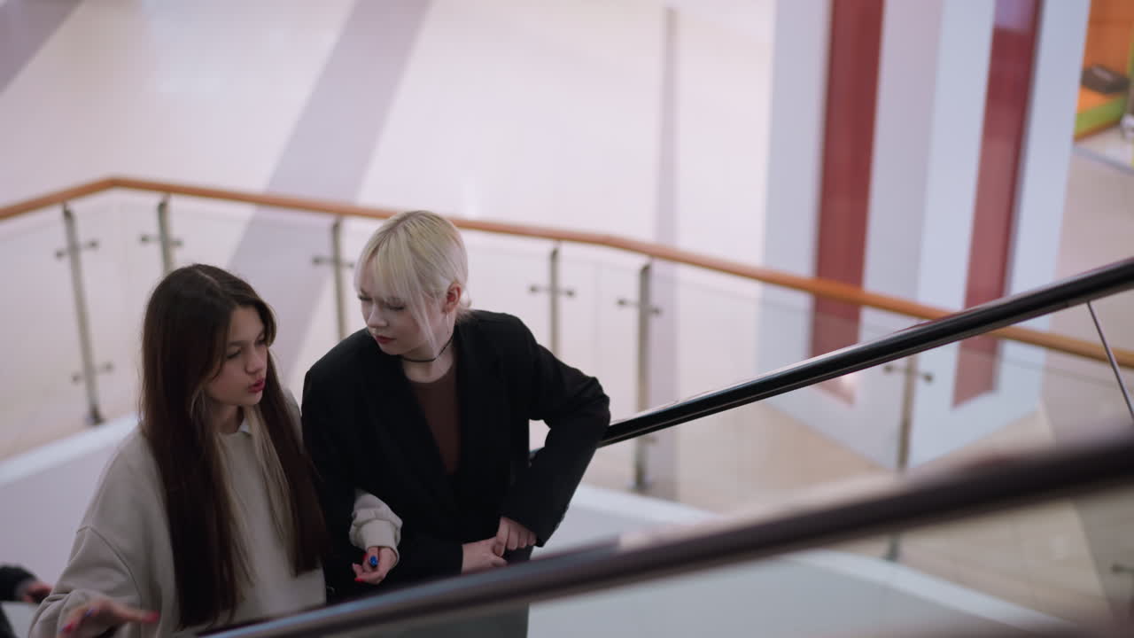 Two friends ride escalator indoors as blonde woman looks with concern at worried companion, scene framed with modern railing, empty tiled floor, and bright shopping mall