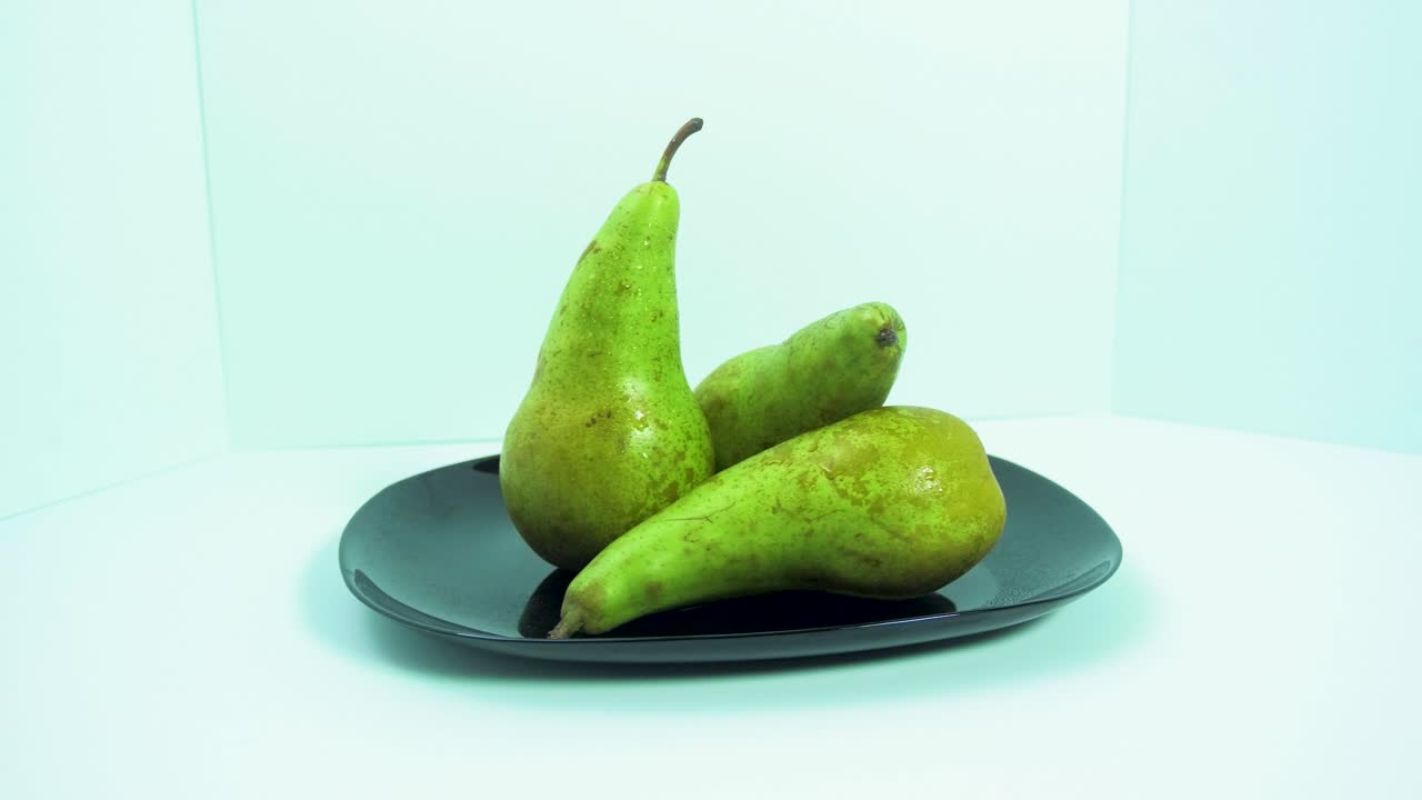 grandes peras verdes frescas giran lentamente sobre un plato negro sobre fondo azul claro, concepto de comida saludable, plano medio, cámara gira a la izquierda