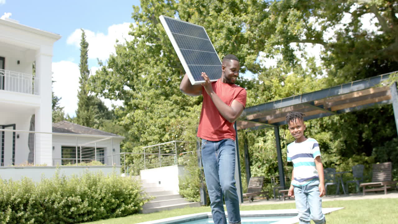 hombre afroamericano sostiene un panel solar, con un niño mirando en un patio trasero soleado en casa