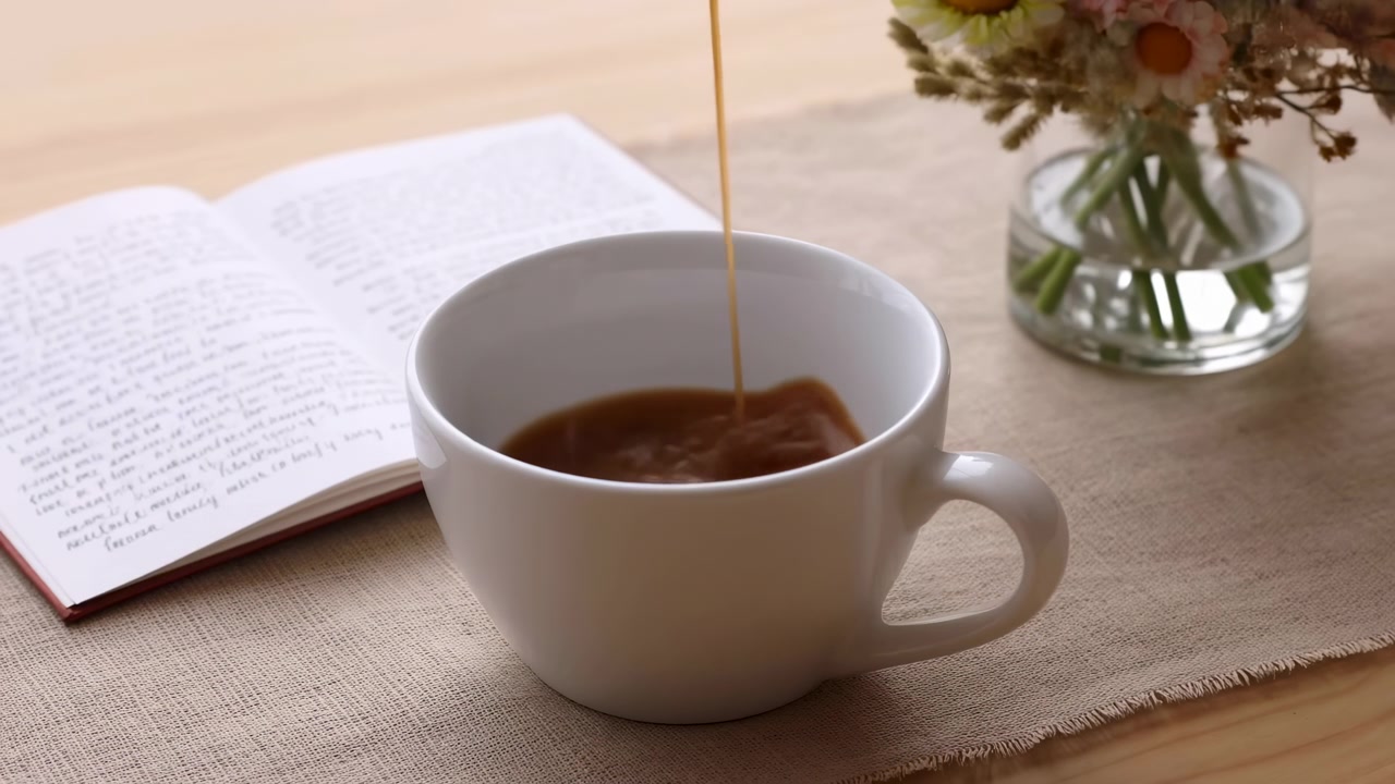 Caffè scuro scorre in una tazza di ceramica bianca su un tavolo coperto di lino, con un libro aperto sulla sinistra e un piccolo vaso di vetro con fiori delicatamente illuminato sullo sfondo.
