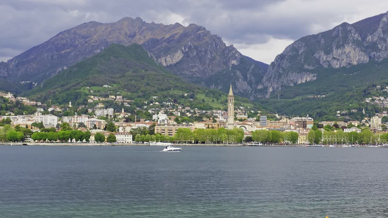 A speedboat is going past a small town on Lake Como with mountains in the background. There are clouds on the mountain tops.