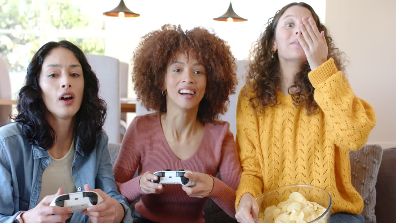 Un grupo diverso de amigas reacciona a momentos cruciales del juego sujetando mandos y comiendo patatas fritas en el sofá