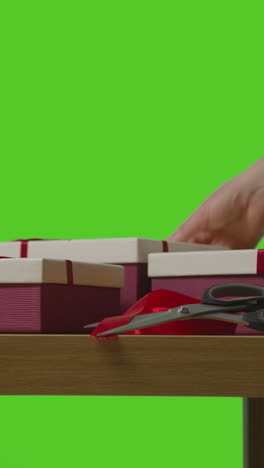 vertical video of man gift wrapping presents in boxes decorated with ribbon on table shot against green screen 1