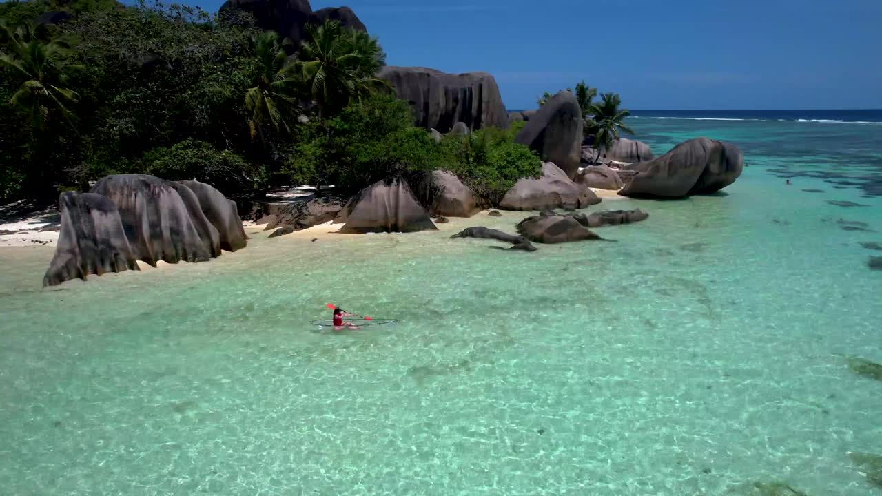 Donna in kayak con fondo trasparente che galleggia accanto alla pittoresca spiaggia tropicale delle Seychelles, Anse Source D'argent, con palma e massi.