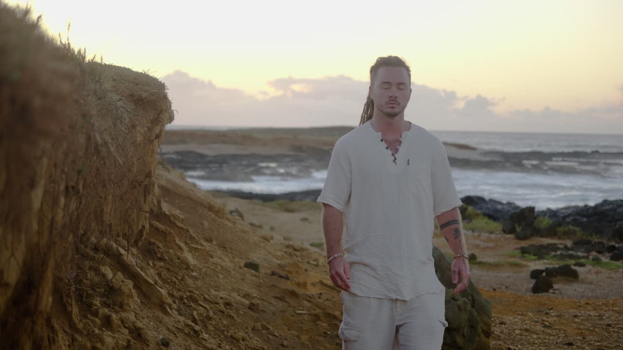 A man in casual white clothing walks toward the camera along a rugged coastal path, with waves breaking against rocks under soft golden sunrise light