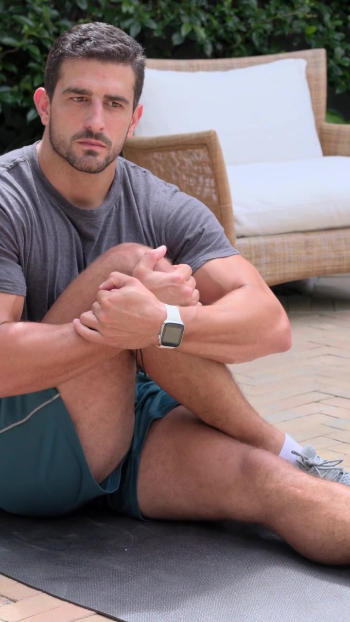 Video verticale: Uomo in maglietta che inizia sdraiato e tira il ginocchio destro per allungare l'anca su un tappetino scuro, nel patio.