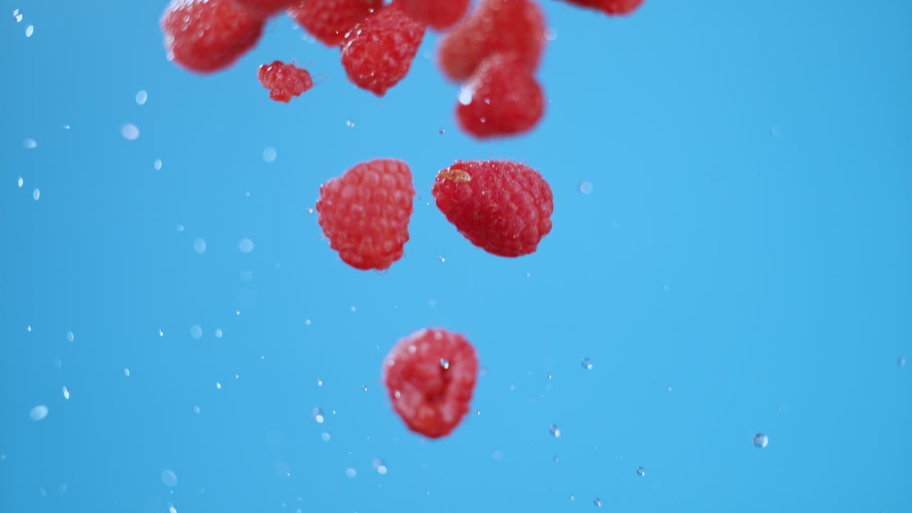 A tight pack of raspberries flying in air with an explosion of water droplets. High quality cinematic studio commercial super slow motion 1000fps shot of berries moving up, down on a blue background.