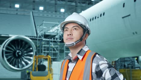 close up side view of asian male engineer with safety helmet with aircraft in the hangar. standing with arms akimbo looking around, checking