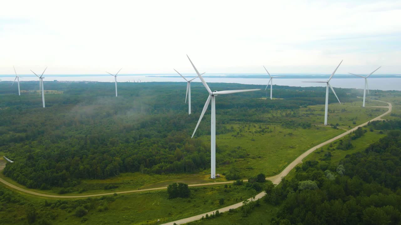 Aerial drone revealing wide shot of large white colored green electrical energy creating wind turbines on a foggy and misty peninsula during a cloudy day spinning and rotating in the wind, Baltic sea