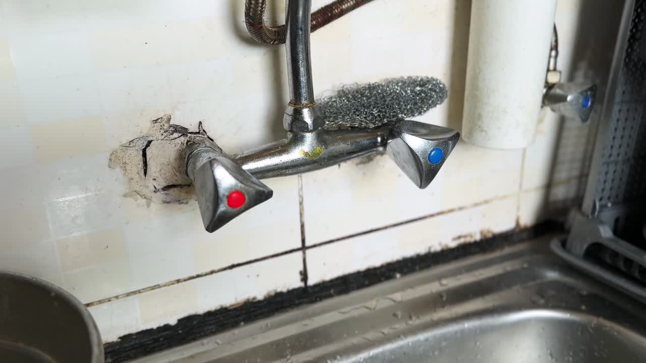 Close-up of a leaking kitchen faucet with hot and cold handles, rusty pipes, and mold buildup on tiled wall above stainless steel sink. Indoor plumbing maintenance and household repair concept