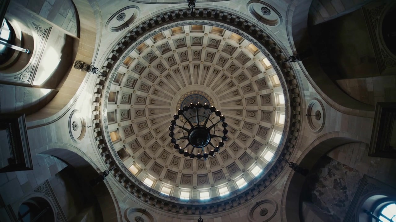 Telecamera che si inclina rivelando una cupola a cassettoni in una rotonda, inquadrando un lampadario, un anello luminoso, archi e murales.