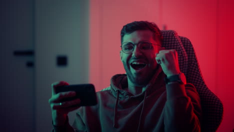 a young man with glasses a professional gamer on a mobile phone plays games and emotionally rejoices in the victory. emotional young man playing mobile games in a neon light. high quality 4k footage