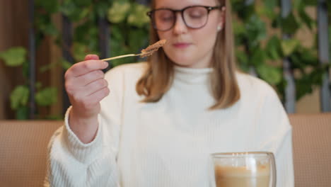 close up of woman in white sweater slicing into chocolate dessert with fork, latte in transparent double-walled cup on side, calm coffeehouse background