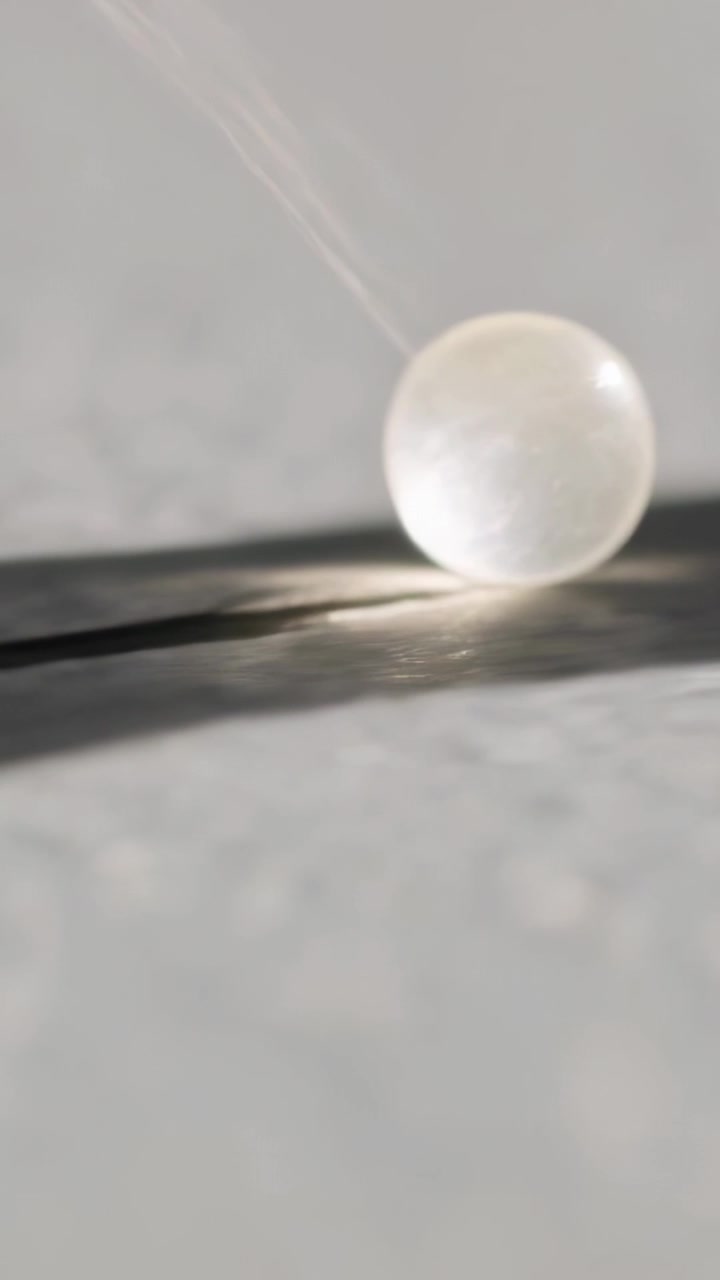 Video verticale: Abbassamento di una perla trasparente su un filo, che tocca del nastro scuro su carta, mostrando un riflesso.