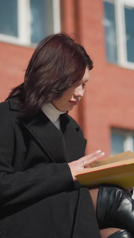 estudiante con abrigo negro y botas de cuero sentada afuera, concentrándose en leer su libro mientras pasa a una nueva página, el cabello revoloteando en el viento con un edificio de ladrillo rojo en el fondo