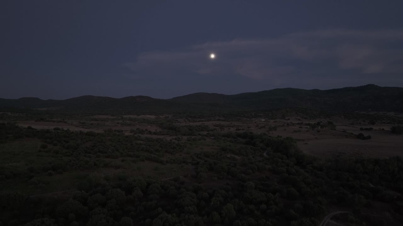 ascending flight with a drone at dusk, a group of trees appears, the field and we see the line of mountains in the background, in the sky the moon appears behind some clouds, it is fantastic