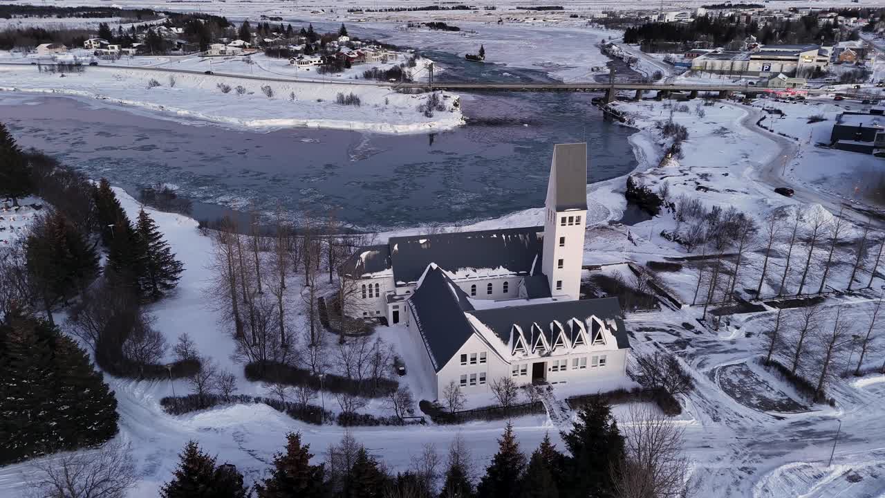 Aerial backwards shot of historic church of Selfoss City in winter. Snowy winter landscape in Iceland. Cars on bridge and flowing ice floe on river. Wide shot.