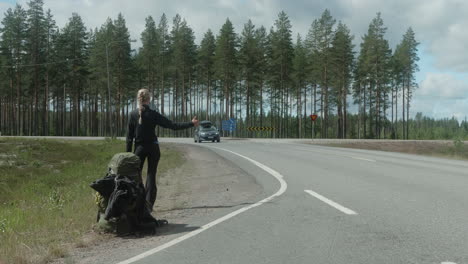 blonde girl hitch hiking on a cross road, finland countryside, female autostopper with heavy backpack in scandinavia, car passing by, wide shot