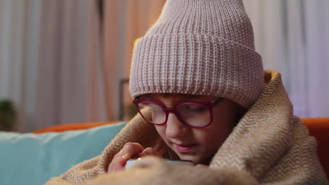 sick toddler girl wear hat wrapped in plaid sit alone shivering from cold on sofa drinking hot cacao