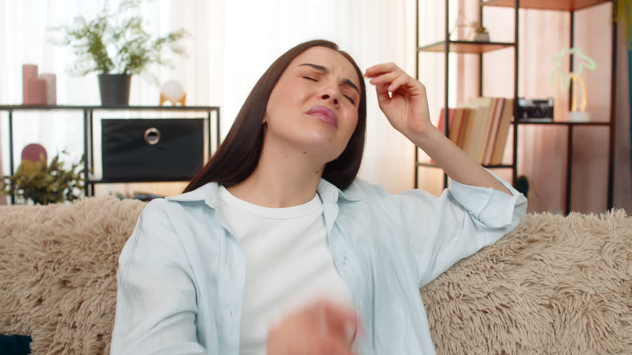 Young woman sitting on sofa holding head suffering from strong headache feeling uncomfortable pain