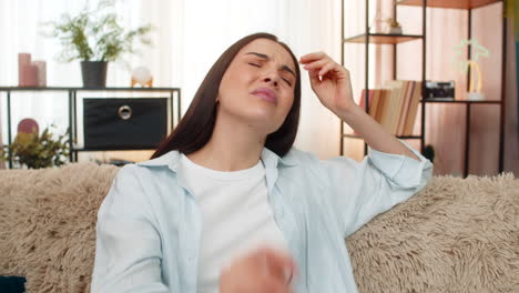 young woman sitting on sofa holding head suffering from strong headache feeling uncomfortable pain