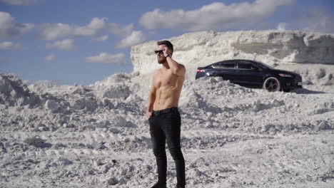 shirtless athlete on white canyon. portrait of handsome man with trained body in the mountains under the blue sky. black car on the hill at sunny day.