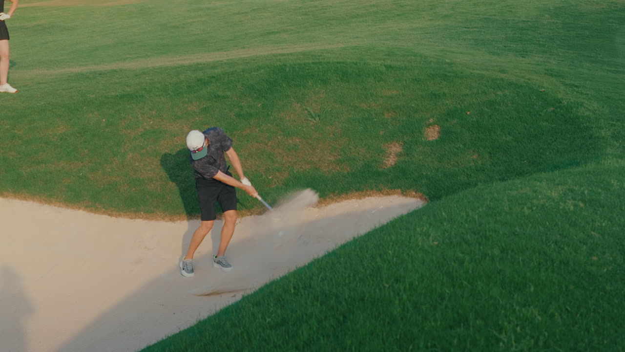 Handheld shot of a male golfer hitting from a greenside bunker in slow motion as his playing partner watches from nearby. Sand flies as the ball lifts, capturing a focused short game moment.