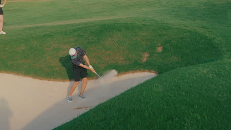 handheld shot of a male golfer hitting from a greenside bunker in slow motion as his playing partner watches from nearby. sand flies as the ball lifts, capturing a focused short game moment.