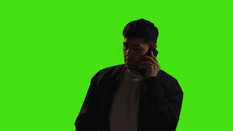 Young-Man-Making-A-Call-On-Mobile-Phone-Standing-Against-Green-Screen-Studio-Background-With-Low-Key-Lighting-1