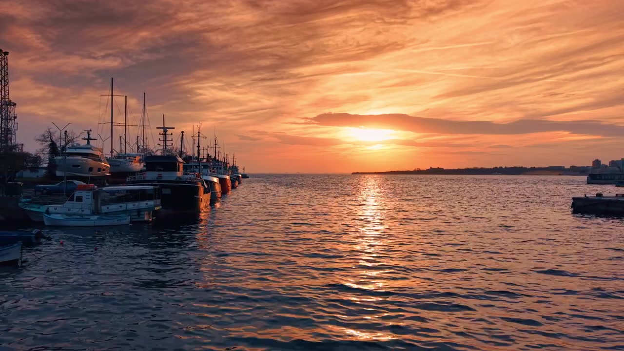 Tramonto su pescherecci ormeggiati al molo del porto. Calda luce dorata si riflette sull'acqua increspata accanto a imbarcazioni e alberi in silhouette sotto un cielo arancione.