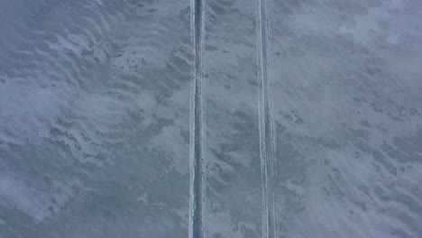 Antenne-Von-Oben-Nach-Unten,-Die-Rückwärts-über-Schneemobilspuren-Im-Matschigen-Schnee-Auf-Einem-Zugefrorenen-See-Fliegt