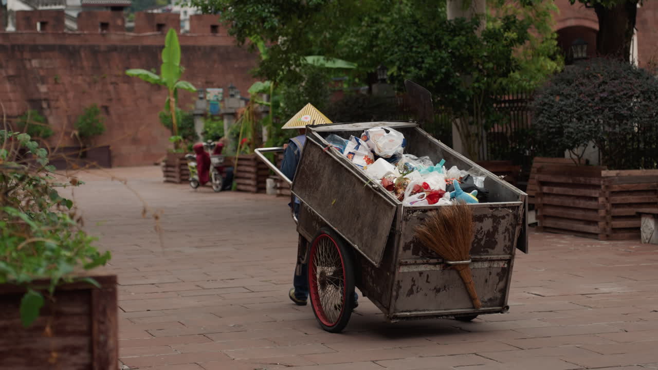 Raccoglitore di rifiuti che spinge un carretto carico attraverso un cortile in mattoni, pieno di sacchi di plastica e vegetazione, con un cappello conico. Carretto di metallo rustico con ruota arrugginita, bancarelle di venditori sullo sfondo, mattina tranquilla.